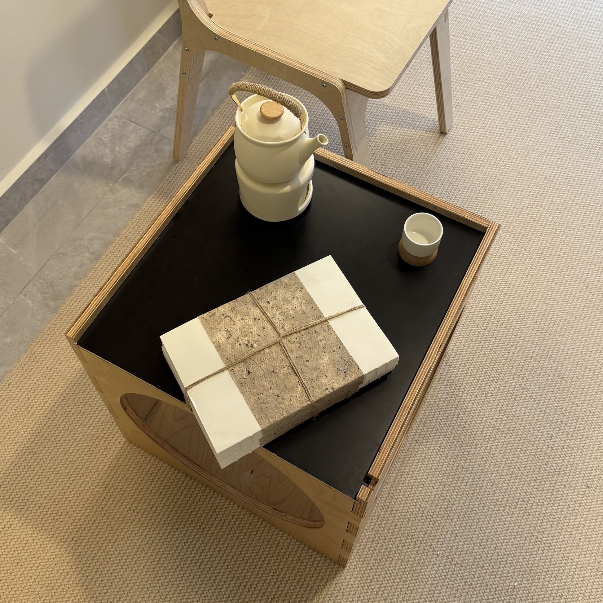 Wooden coffee table with black top displaying a teapot and cups, next to a chair on a carpeted floor.
